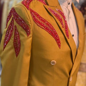 Könnte beinhalten: Senfgelber Blazer mit roten Perlenverzierungen auf den Schultern. Die zweireihige Jacke hat ein fallendes Revers und zwei Knöpfe. Ein weißes Hemd ist darunter zu sehen.
