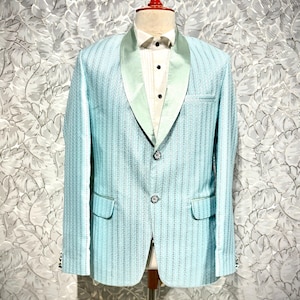 May include: A light blue blazer with a satin lapel and two front pockets. The blazer has a textured pattern and is displayed on a mannequin. The outfit includes a white dress shirt and a bow tie.