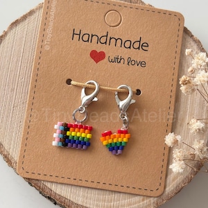 Op de afbeelding: Twee bedelarmbanden met zilveren sluitingen. De ene bedel is een regenboogvlag, de andere een regenbooghart. De bedels zijn bevestigd aan een bruine kaart met de tekst "Handmade with love". De achtergrond is een houten schijf.