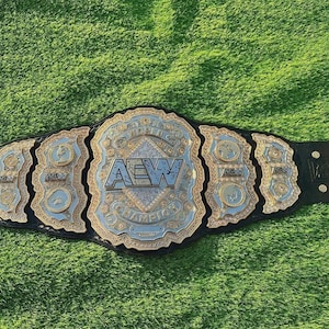 May include: A wrestling championship belt with a black leather strap and a large, ornate center plate. The center plate features the letters "AEW" in silver, surrounded by gold and blue detailing. The belt is set against a green background.