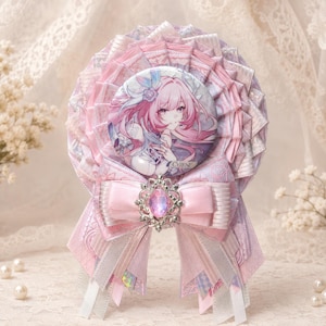 May include: A circular rosette badge featuring a central pinback button with an anime-style illustration of a character with long pink hair. The rosette is constructed from layered pink and white ribbons with a central silver-toned brooch with a pink gem.