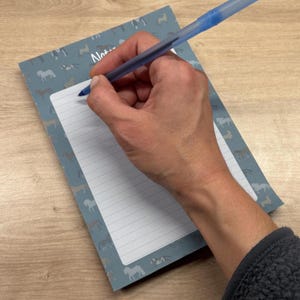 May include: A blue notepad featuring a repeating pattern of light grey horse silhouettes and the word Notes printed at the top, accompanied by a translucent blue ballpoint pen positioned over the lined white writing area of the paper.