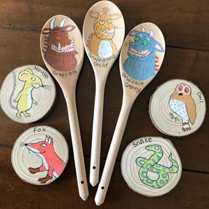 May include: Three wooden spoons and four wooden slices painted with characters from The Gruffalo. The spoons feature the Gruffalo, Gruffalo's Child, and Gruffalo Granny. The slices depict a mouse, fox, owl, and snake with their names written.