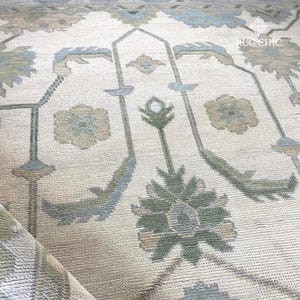 May include: Close-up of a beige rug with a floral pattern in shades of blue, green, and tan. The rug has a soft, textured appearance and a rolled edge. The design includes stylized floral and geometric elements.