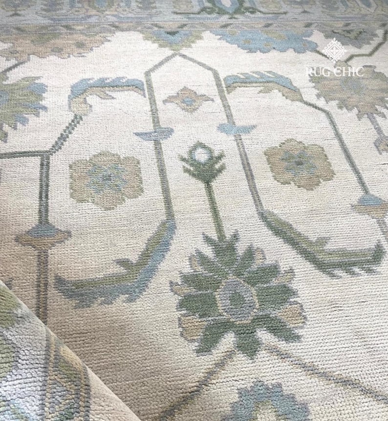 May include: Close-up of a beige rug with a floral pattern in shades of blue, green, and tan. The rug has a soft, textured appearance and a rolled edge. The design includes stylized floral and geometric elements.