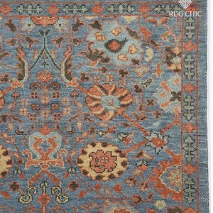 Op de afbeelding: Detail van een blauw tapijt met een ingewikkeld bloemenpatroon in tinten oranje, beige en bruin. Het tapijt heeft een gefranjerde rand en de tekst "RUG CHIC" is rechtsboven zichtbaar.