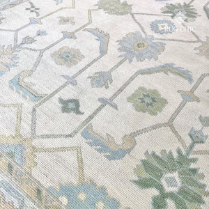May include: Close-up of a cream-colored rug with an intricate floral pattern. The design features stylized flowers and geometric shapes in shades of blue, green, and beige. The rug has a textured appearance, suggesting a woven construction.