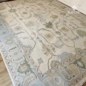 May include: A large, rectangular area rug with a cream-colored background and an intricate floral pattern in shades of green, blue, and beige. The rug has a detailed border with geometric and floral designs, adding a touch of elegance to the floor covering.