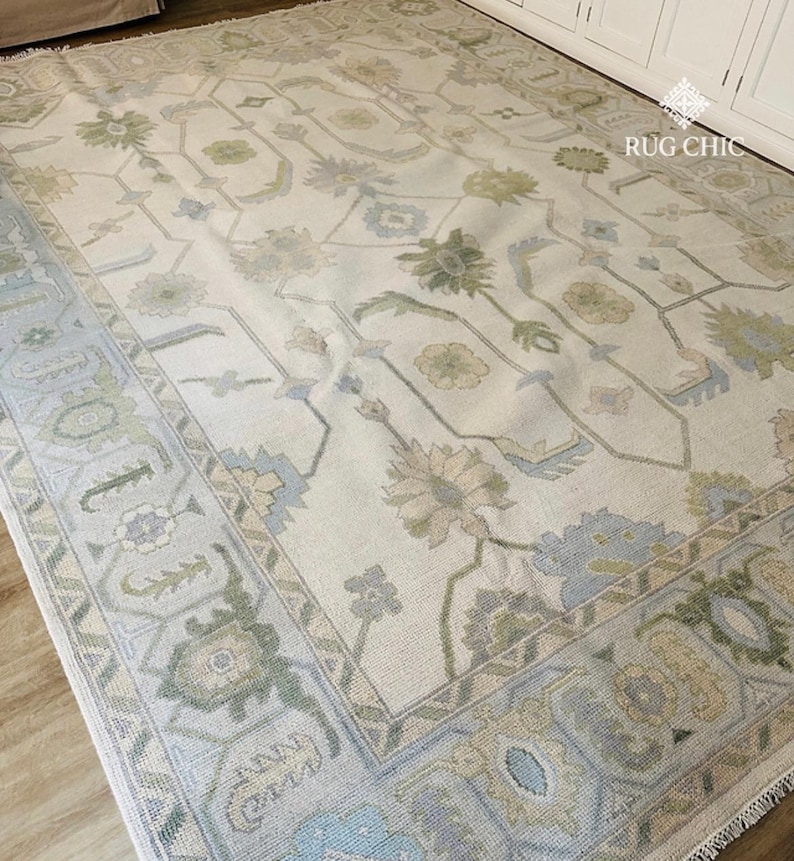 May include: A large, rectangular area rug with a cream-colored background and an intricate floral pattern in shades of green, blue, and beige. The rug has a detailed border with geometric and floral designs, adding a touch of elegance to the floor covering.