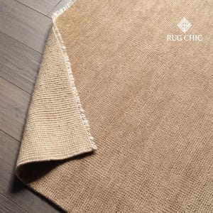 May include: A beige woven rug with a textured surface. The corner of the rug is folded over, revealing the underside. The rug has a fringe edge and the text "RUG CHIC" is visible.