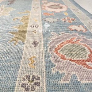 May include: Close-up of a light blue rug with a detailed pattern. The rug features floral and geometric designs in shades of pink, orange, green, and purple. The text "RUG CHIC" is visible on the rug.