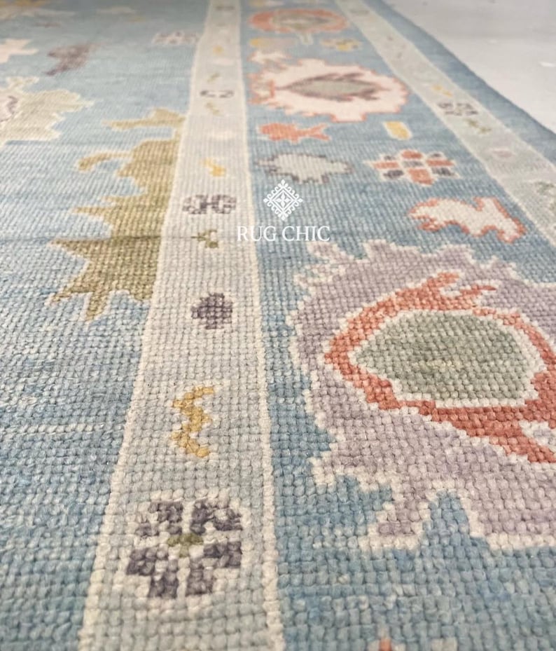 May include: Close-up of a light blue rug with a detailed pattern. The rug features floral and geometric designs in shades of pink, orange, green, and purple. The text "RUG CHIC" is visible on the rug.
