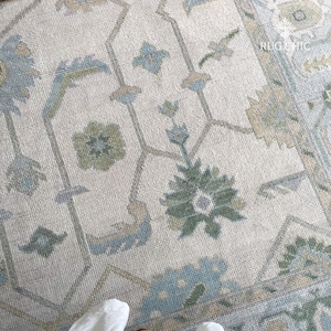 May include: Close-up of a beige rug with an intricate floral and geometric pattern in shades of blue, green, and tan. The rug has a distressed, vintage look, with the text "RUG CHIC" visible in the upper right corner.