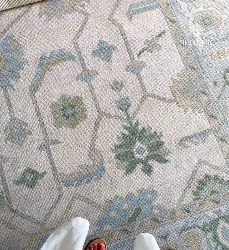 May include: Close-up of a beige rug with an intricate floral and geometric pattern in shades of blue, green, and tan. The rug has a distressed, vintage look, with the text "RUG CHIC" visible in the upper right corner.