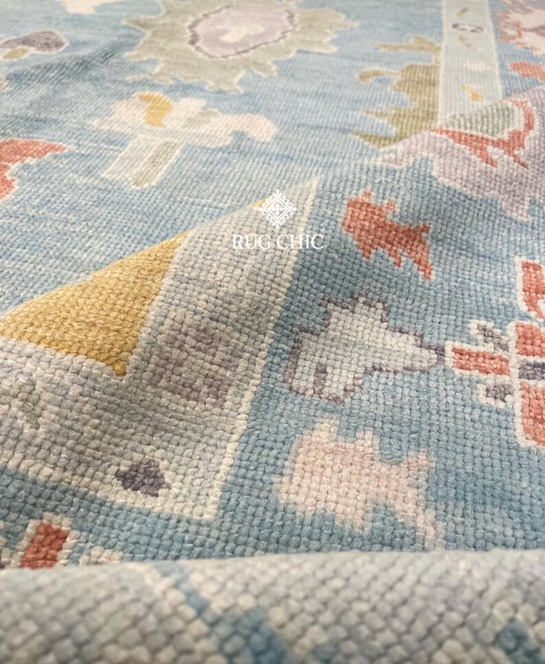 May include: Close-up of a light blue rug with a detailed pattern. The rug features a mix of colors, including peach, yellow, and gray, creating a textured appearance. The text "RUG CHIC" is visible on the rug.