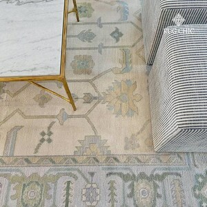 May include: An overhead shot of a cream-colored rug with a detailed floral pattern in shades of blue, green, and beige. A gold-legged coffee table with a marble top and a striped ottoman are partially visible.