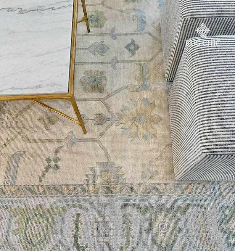 May include: An overhead shot of a cream-colored rug with a detailed floral pattern in shades of blue, green, and beige. A gold-legged coffee table with a marble top and a striped ottoman are partially visible.