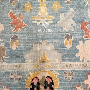 May include: A blue rug with a repeating pattern of abstract shapes in shades of orange, yellow, gray, and white. The rug has a border design and the text "RUG CHIC" is visible. Black sandals are visible at the bottom of the image.