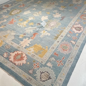 May include: A large, rectangular area rug with a light blue background and a detailed border. The rug features a central design with floral and abstract patterns in shades of pink, yellow, and gray. The text "RUG CHIC" is visible on the rug.