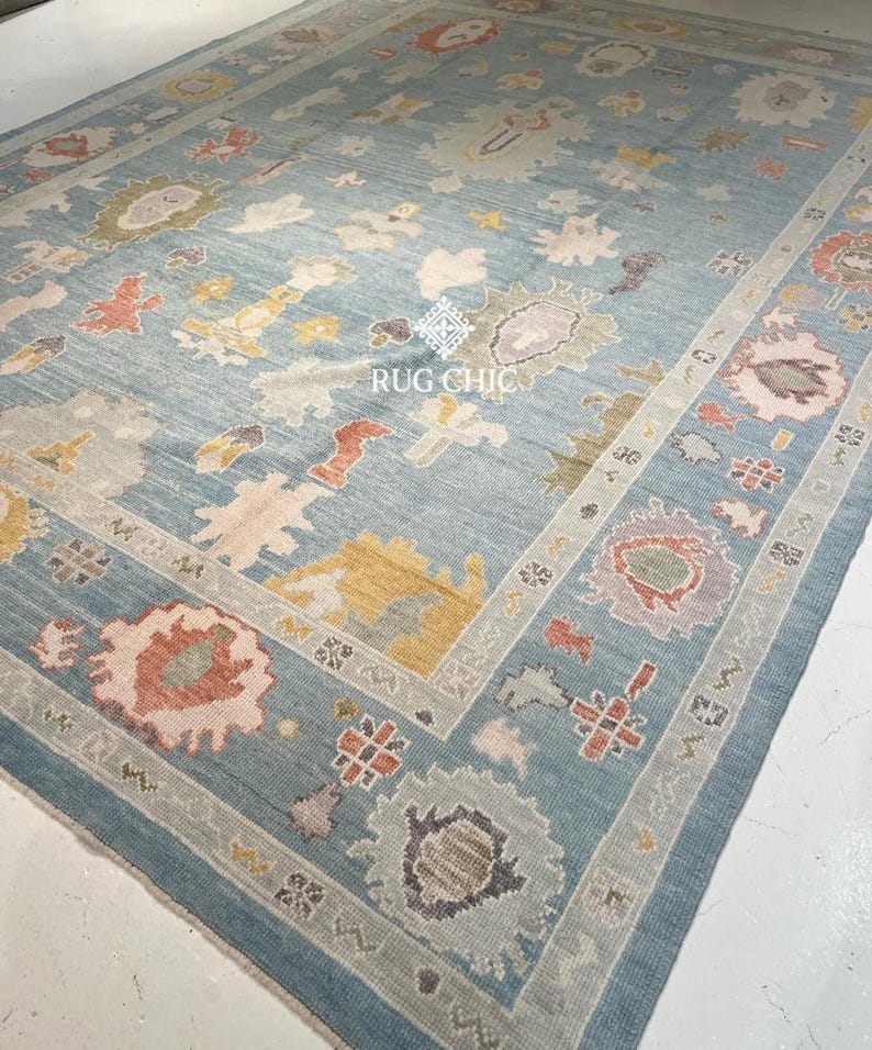 May include: A large, rectangular area rug with a light blue background and a detailed border. The rug features a central design with floral and abstract patterns in shades of pink, yellow, and gray. The text "RUG CHIC" is visible on the rug.