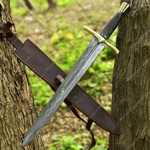 May include: A medieval sword with a patterned blade and a gold-coloured crossguard. The handle is wrapped in a dark, textured material. The sword rests in a brown leather sheath, leaning against a tree.