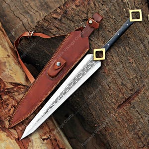 May include: A decorative sword with a silver blade featuring an etched floral design. The sword has a black handle with gold-coloured square accents and rests in a brown leather sheath with a shoulder strap. The sheath is stitched with a decorative pattern.