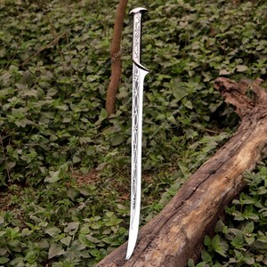以下が含まれることがあります： 装飾的な柄と刃のデザインが施された銀色の剣。刃は湾曲したエッジと装飾的な切り抜きがあります。剣は屋外で、風化した丸太の上に置かれ、緑の葉を背景に展示されています。