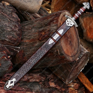 May include: A Viking-style sword with a brown leather sheath, featuring intricate silver-colored Celtic knotwork and decorative metal accents. The handle is wrapped in brown leather. The sword rests on a pile of wooden logs.