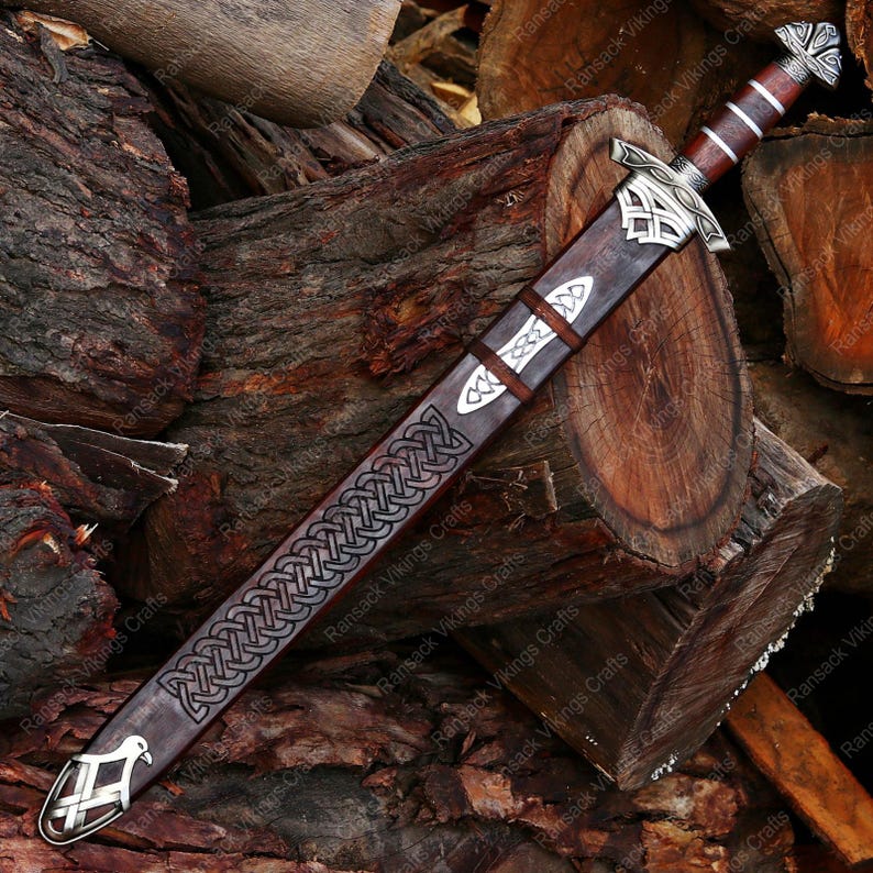 May include: A Viking-style sword with a brown leather sheath, featuring intricate silver-colored Celtic knotwork and decorative metal accents. The handle is wrapped in brown leather. The sword rests on a pile of wooden logs.