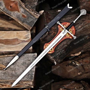 May include: A silver sword with an ornate handle and a long, sharp blade, displayed on a wooden plaque. A black sheath is positioned to the left. The sword is set against a backdrop of stacked wooden logs.