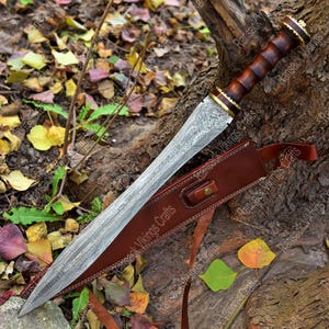 May include: A Damascus steel sword with a wooden handle and a brown leather sheath. The sword has a detailed pattern on the blade and is resting on a bed of leaves and twigs. The handle is dark brown with gold accents.