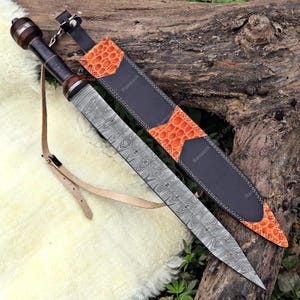 May include: A Damascus steel sword with a wooden handle and leather sheath. The sheath is black with orange patterned accents. A leather strap is attached to the handle. The sword is lying on a wooden surface.