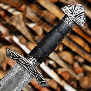 May include: Close-up of a Viking sword with a patterned blade and ornate silver-colored hilt. The handle is black, and the pommel features a detailed design. The sword is set against a blurred wooden background, showcasing the craftsmanship.