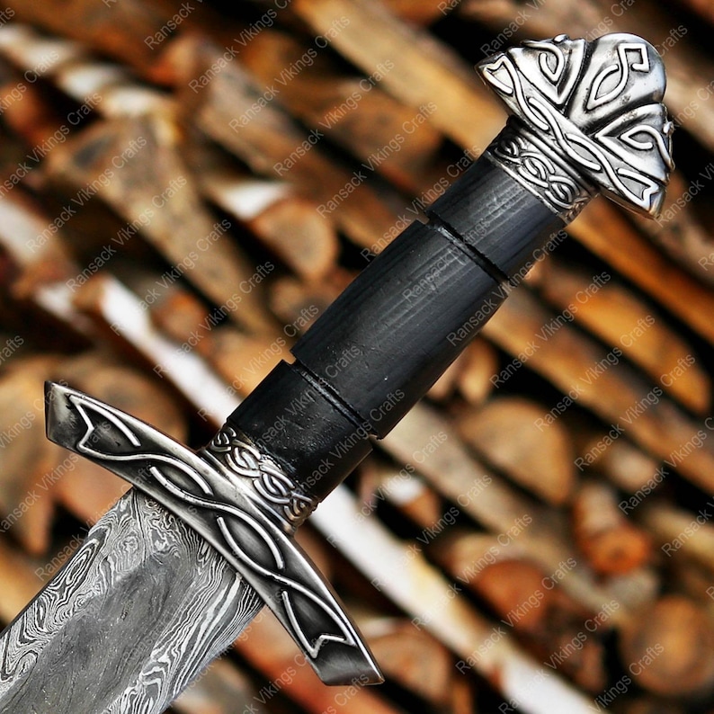 May include: Close-up of a Viking sword with a patterned blade and ornate silver-colored hilt. The handle is black, and the pommel features a detailed design. The sword is set against a blurred wooden background, showcasing the craftsmanship.