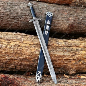 May include: A Viking sword with a patterned blade and a black handle, resting on a wooden surface. The sword is accompanied by a black sheath with silver accents. The handle features intricate silver detailing.