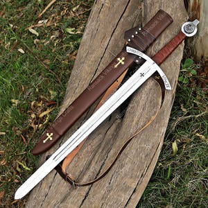 May include: A medieval sword with a silver blade and a brown leather-wrapped handle, displayed on a wooden surface. The sword has a brown leather sheath and a leather strap. The blade is inscribed with "CHRISTUS" and "VINCIT".