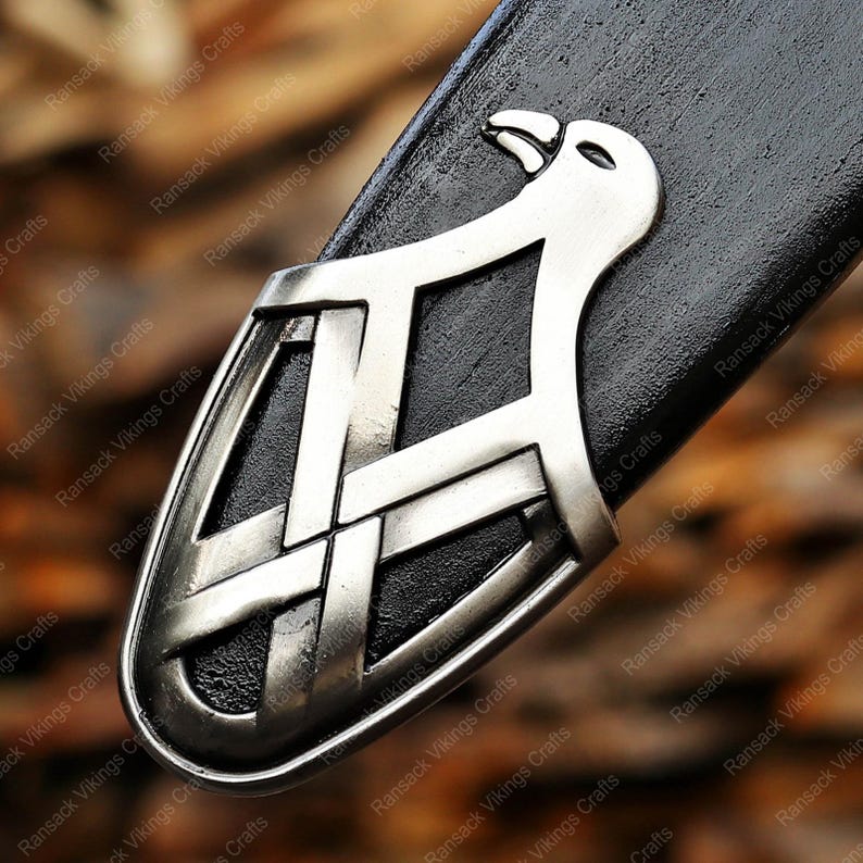 May include: Close-up of a silver-colored metal sword pommel with an intricate design. The pommel features a stylized bird and interwoven patterns, set against a black background. The image is from Ransack Vikings Crafts.