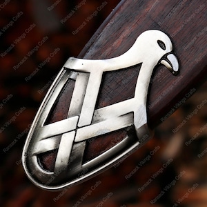 May include: Close-up of a silver-colored metal sword guard with a bird-like design. The guard is mounted on a dark wooden surface. The design features a bird's head and a geometric pattern. The text "Ransack Vikings Crafts" is visible.