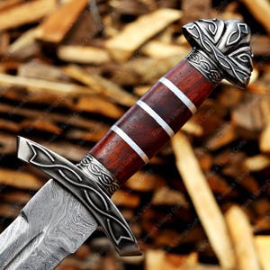 May include: Close-up of a decorative sword with a patterned blade and a wooden handle. The handle is dark brown with silver accents and a detailed silver pommel. The blade has a unique pattern.