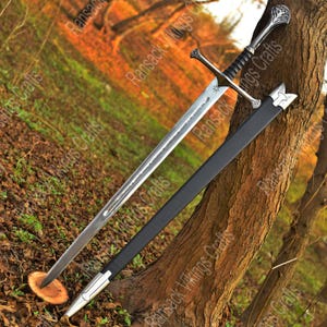 May include: A long, silver sword with a black handle and crossguard, in a black sheath. The sword features detailed hilt and crossguard designs. The blade is etched with a pattern. The sword is propped against a tree trunk.