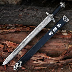 May include: A detailed image of a sword with a patterned blade and a black handle. The sword is accompanied by a black sheath with silver accents. The design features intricate metalwork and is set against a rustic wooden background.