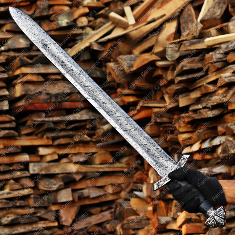 May include: A long, silver sword with an ornate handle and blade. The blade features a detailed pattern. The sword is held by a person wearing black gloves. The background is a stack of wooden logs.