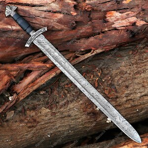 May include: A long, silver sword with a patterned blade and a black handle. The sword features a decorative crossguard and pommel. The blade is approximately 30 inches long. The sword is displayed on a wooden surface.