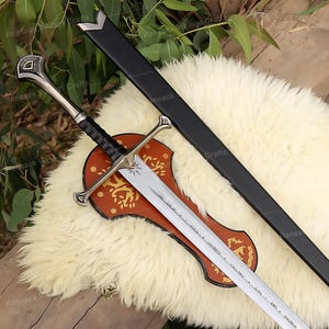 May include: A silver and black sword with a black sheath. The sword has a detailed hilt and crossguard, with a black handle. The blade is engraved with text and is displayed on a decorative wooden plaque. The sword rests on a white fur-like surface.