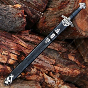 May include: A black sword with a silver hilt and accents, resting on a pile of wood. The sword features intricate designs on the hilt and scabbard, with a black handle and a silver crossguard. The scabbard is black with silver details.