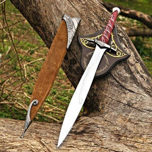 May include: A fantasy sword with a silver blade and a red and white handle, displayed on a brown wooden shield with gold detailing. A brown leather sheath with silver accents is also present. The sword is on a weathered log.