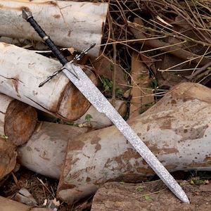 May include: A long, silver sword with a textured blade and a black handle. The sword features a crossguard and a pointed tip. The handle is wrapped in black material. The sword is resting on a pile of logs.