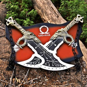 May include: A wall decoration with two crossed blades, each with a dragon head handle and detailed patterns. The blades are black and white, set against a red and black backdrop with the words "GOD OF WAR".