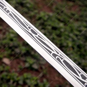 以下が含まれることがあります： 精巧な黒い装飾模様が施された銀色の剣の刃のクローズアップ。 刃は研磨仕上げで、デザインは様式化された形と線が特徴です。 背景はぼやけた緑色です。