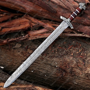 May include: A long, ornate sword with a patterned steel blade and a wooden handle. The handle is dark brown with silver accents. The sword is lying on a bed of brown wood.